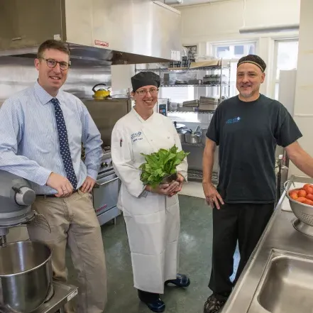 Three members of 餐厅 staff in a kitchen. 一个男人站在一碗西红柿旁边，一个女人拿着一捆蔬菜.